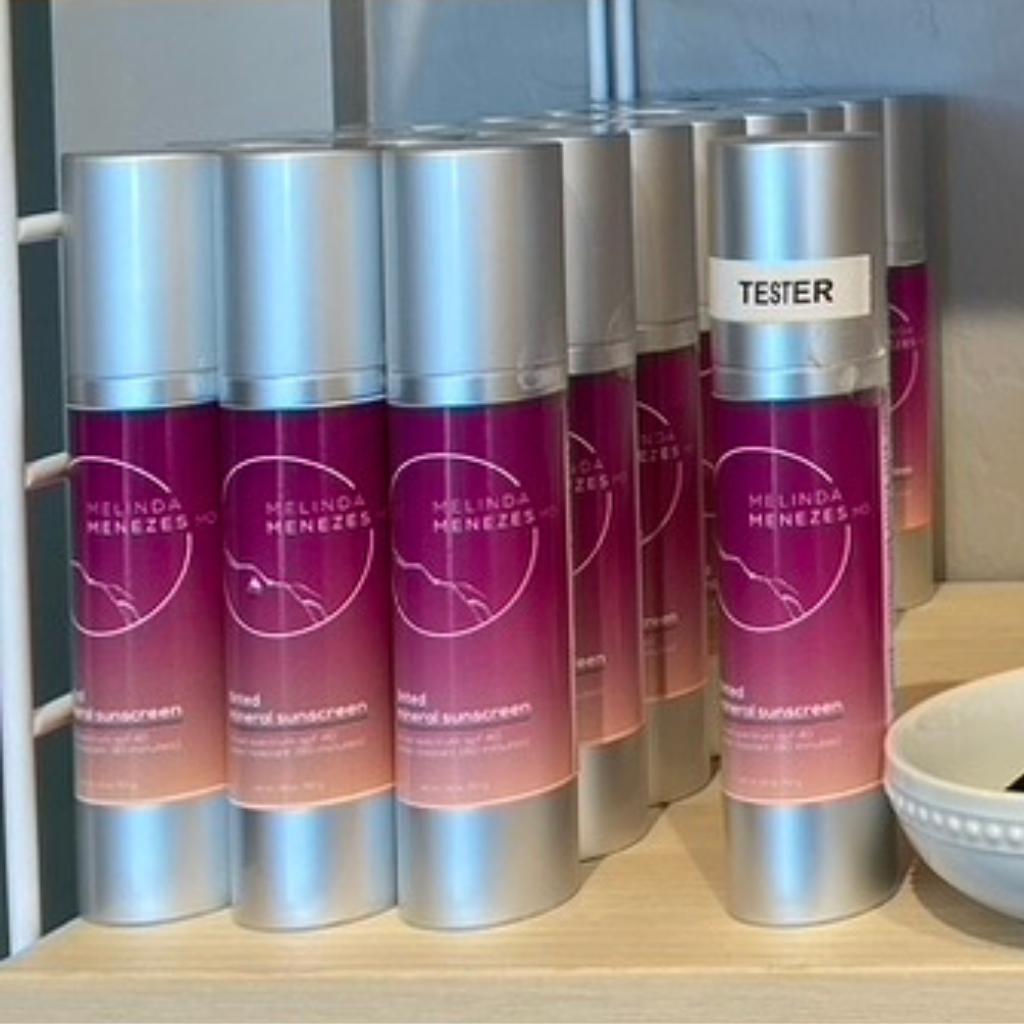 Melinda Menezes MD sunscreen sitting on the shelf in the clinic boutique. The labels are a vibrant magenta color and one bottle is labeled as a tester